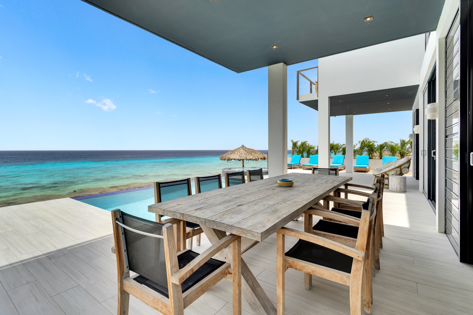 Luxury Bonaire villa with a covered oceanfront dining terrace featuring a rustic wood table, infinity pool overlooking the turquoise Caribbean Sea.